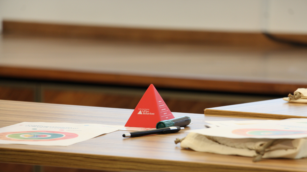 Foto de uma mesa. Em cima, dá para ver um triângulo pequeno vermelho, canetas e papéis.