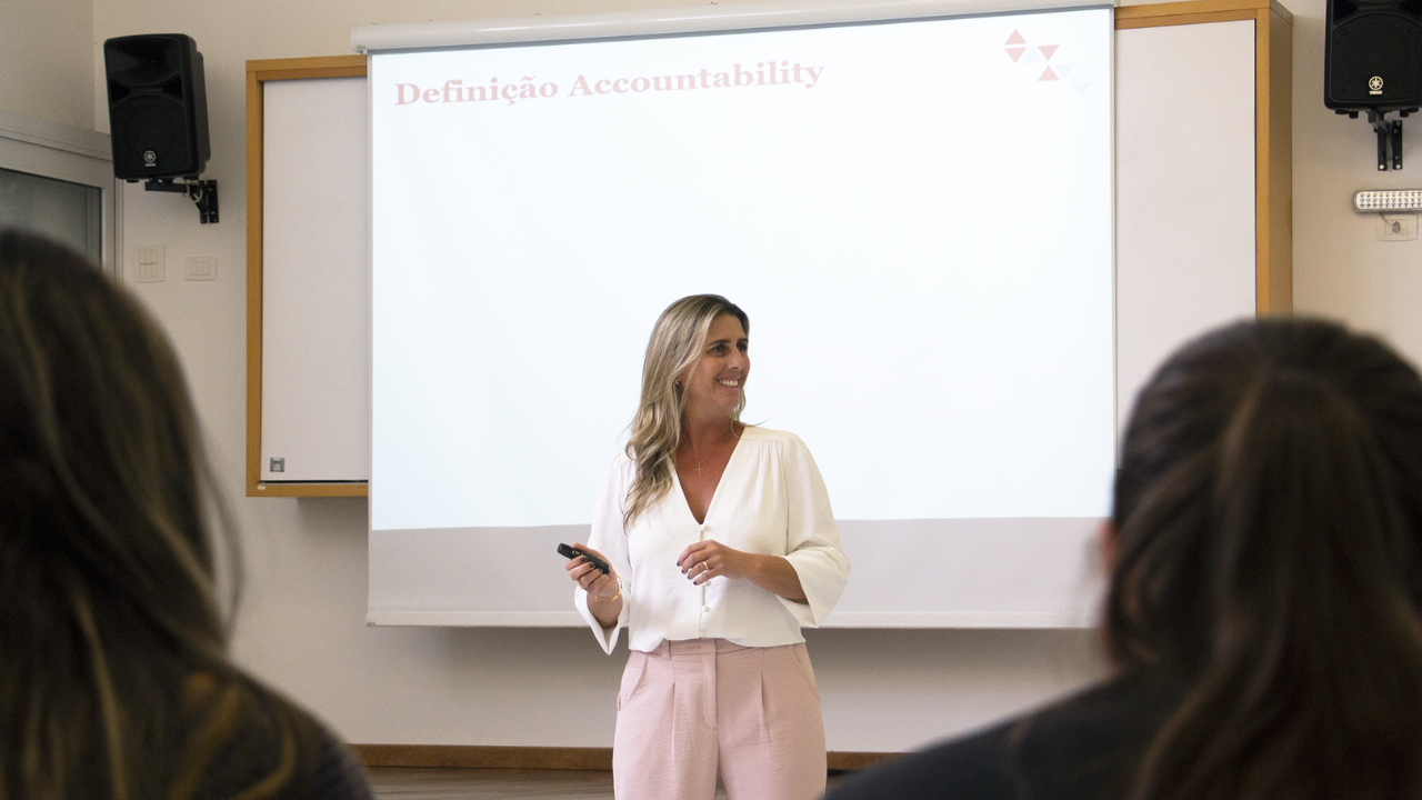 Photo of our inspiring Paula Minetto, in the classroom. Paula is a white woman with blonde hair, wearing a white shirt. In the background, you can see a blank screen.